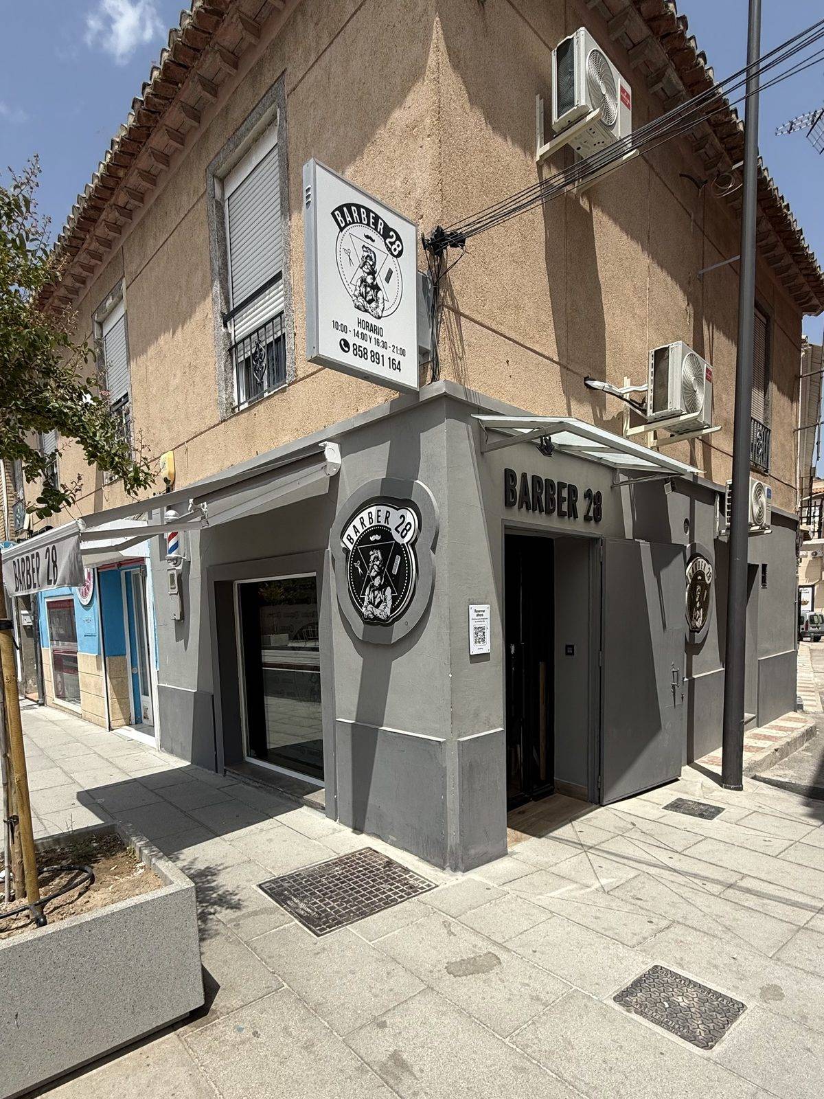 The Churriana de la Vega shop from the street: grey corner with the shop's emblem, the BARBER 28 sign and the opening-hours board overhead
